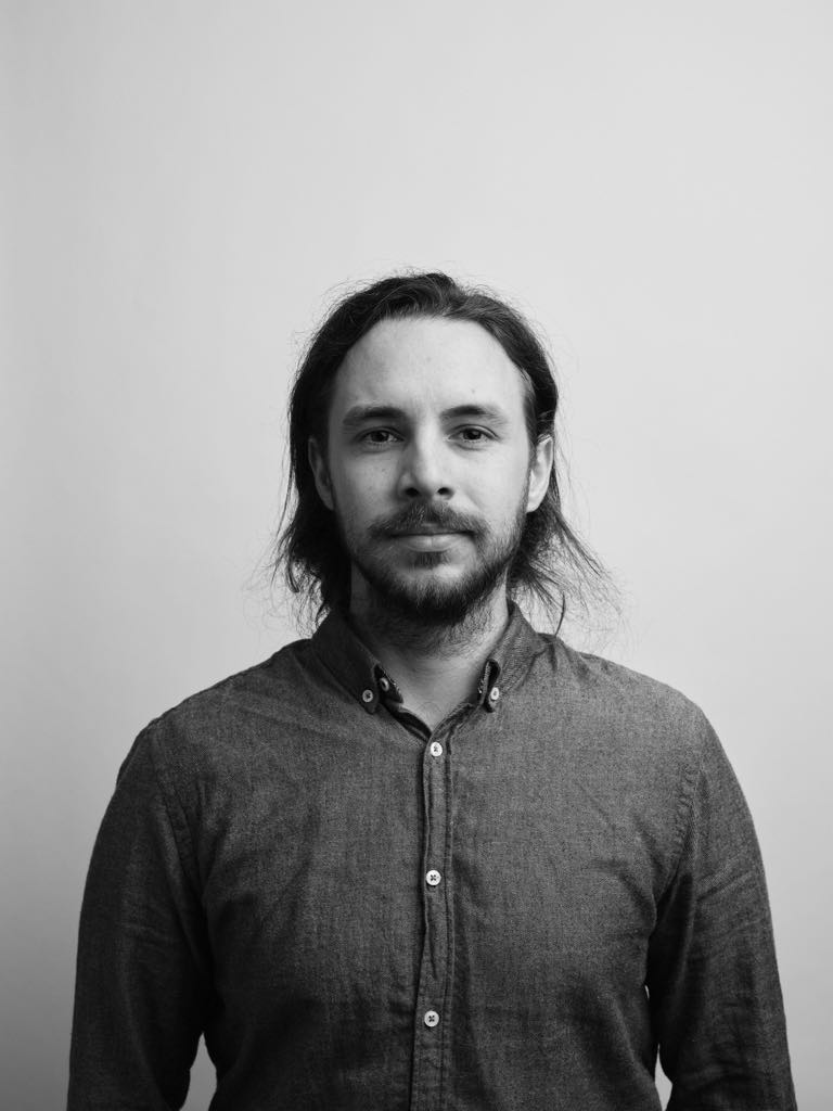 Black and white image of a young man. Long dark hair tied back. Smiling and looking into the camera